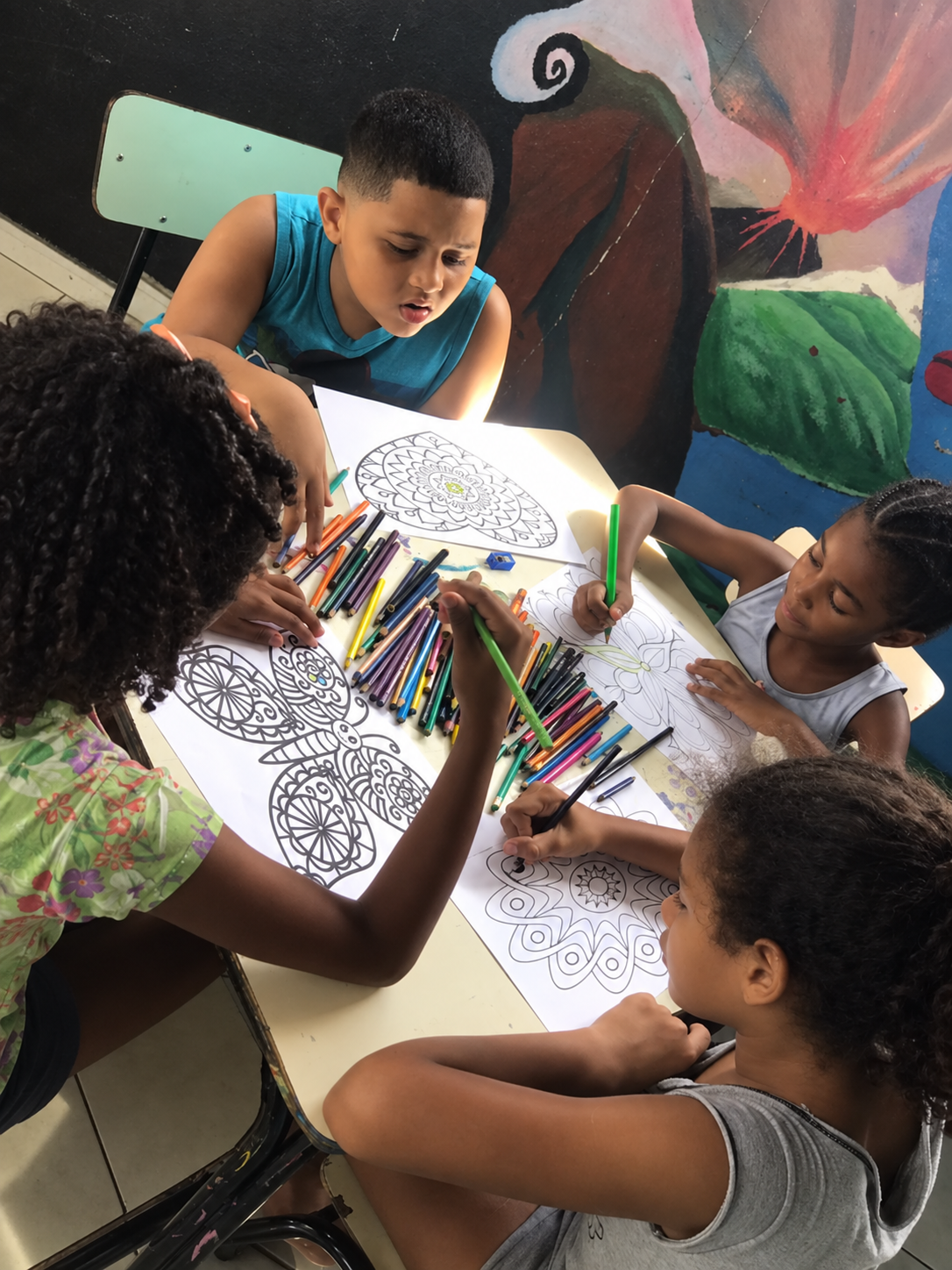 Crianças colorindo desenhos de mandala em mesa coletiva com uma educadora, cercadas de lápis de cor.