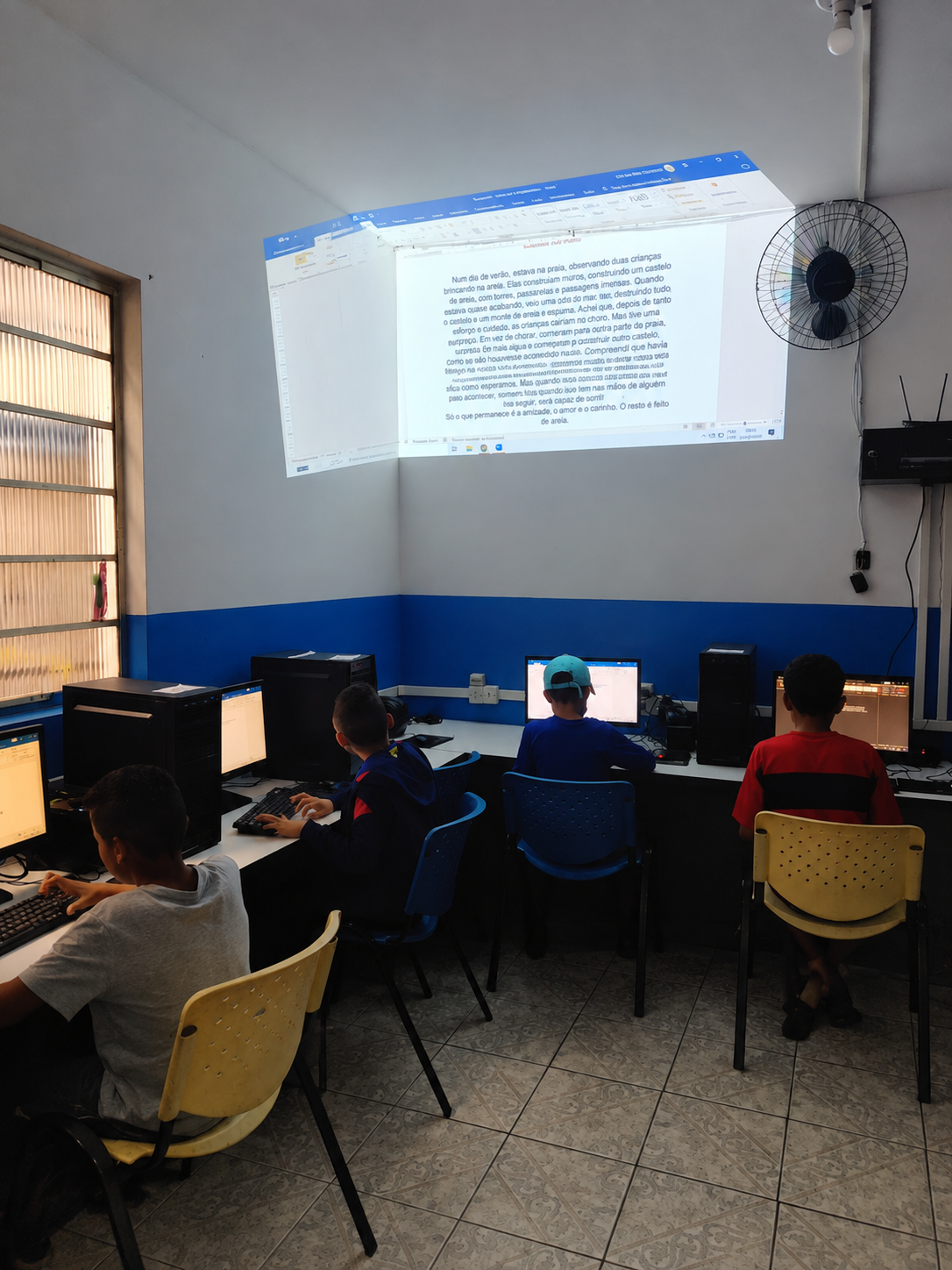 Crianças em laboratório de informática digitando em computadores, com texto de produção escrita projetado na parede
