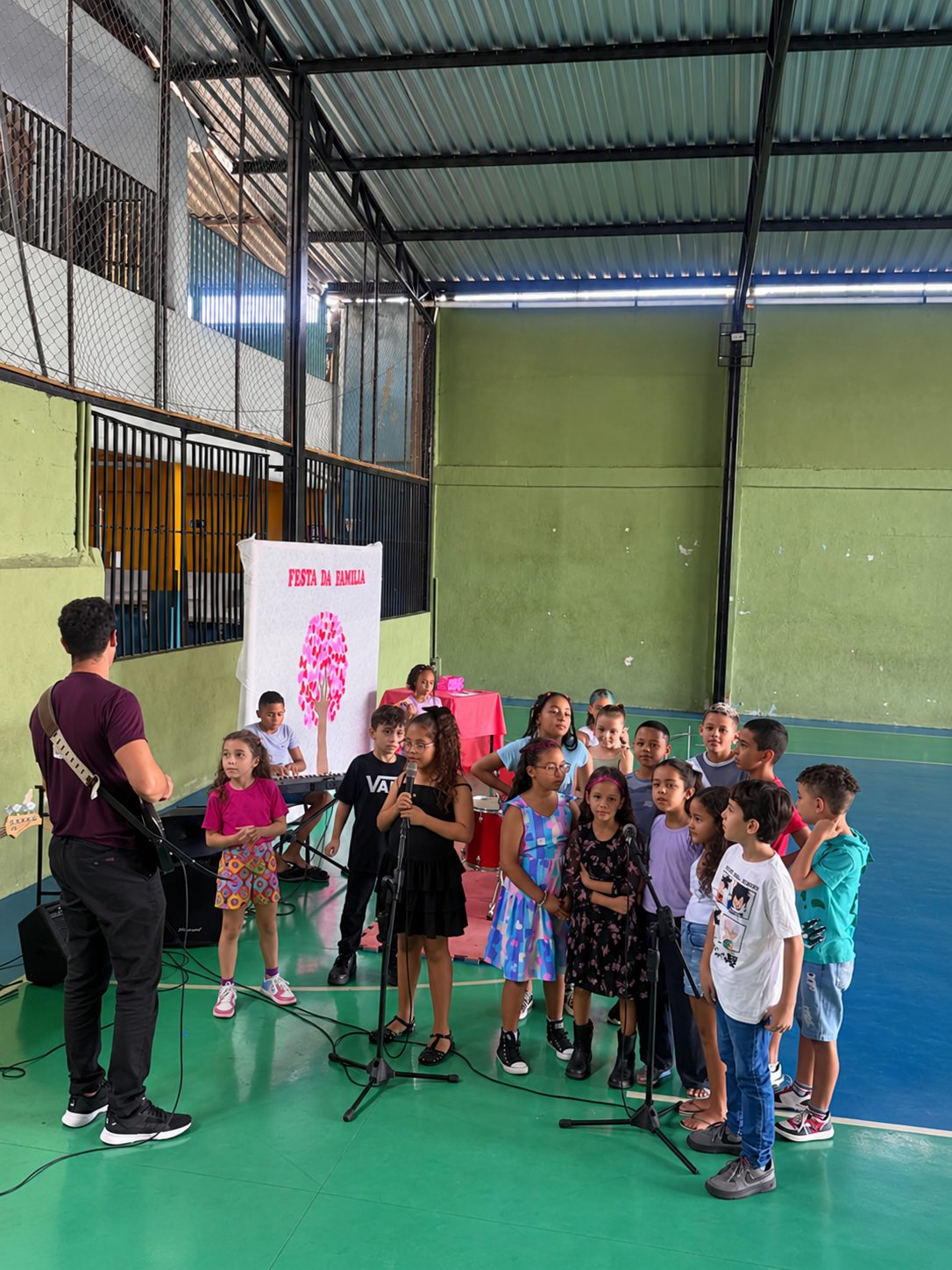 Crianças cantando em microfones ao lado de educador tocando violão, em evento com faixa 'Festa da Família'